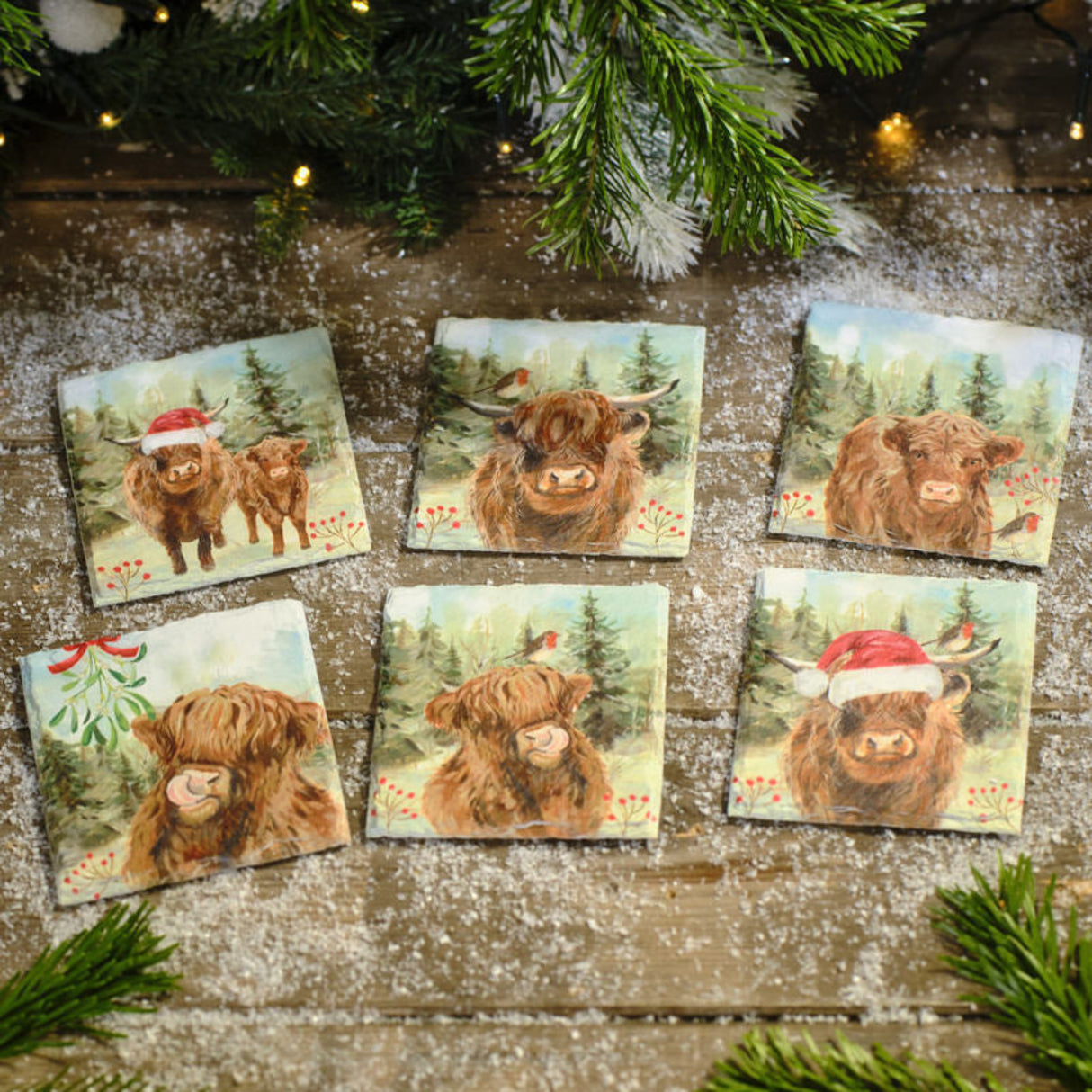 Set of six coasters with highland cow illustrations on a wooden surface with Christmas decorations.