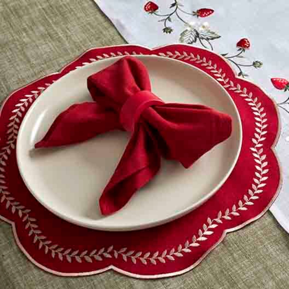 Red folded napkin on a white plate with a decorative red rim, placed on a tablecloth with strawberry pattern.